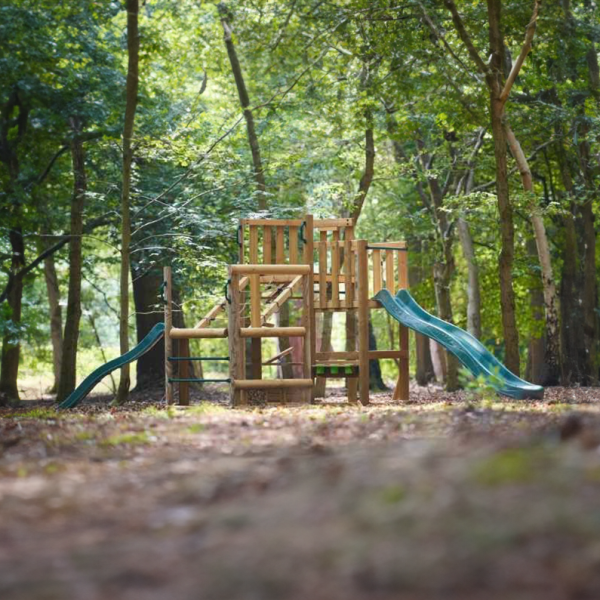 Holz-Spielplatzstruktur mit zwei Rutschen in einem von Bäumen umgebenen Waldgebiet.